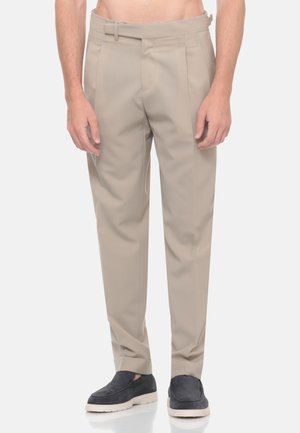 Uomo con pantaloni beige su misura e scarpe slip-on grigio scuro in piedi contro uno sfondo bianco semplice.