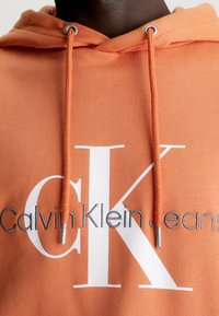 Orange hooded sweatshirt with drawstrings, featuring a large white CK logo and smaller gray "Calvin Klein Jeans" text. Soft fabric texture.