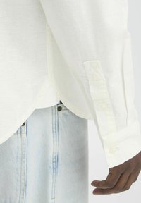 Camisa blanca de manga larga con puños abotonados, hecha de tela ligera. Dobladillo curvado visible por encima de unos vaqueros azules con detalles de botones visibles.