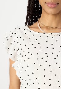 Valkoinen mekko, jossa on mustia pilkkuja, röyhelöhihoja ja teksturoitua kangasta. Täydennetty kerroksilla helmistä ja kultaketjuista koostuvilla kaulakoruilla.