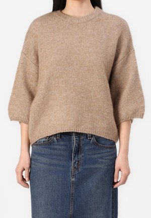 Vrouw die een beige gebreide trui met driekwartmouwen en een blauwe denim rok draagt, staand tegen een effen achtergrond.