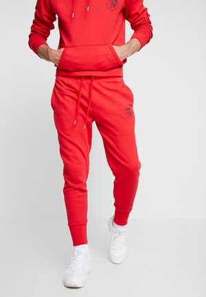 Czerwona bluza z kapturem z przednią kieszenią i logo, połączona z pasującymi dresami z elastyczną talią. Strój ma miękką, gładką fakturę.