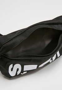 Svart dragkedja på påse med vit fet text, delvis öppen för att visa det tomma inre mot en ljus bakgrund.