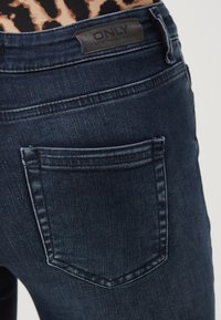 Jeans de mezclilla azul oscuro con detalles de costura, un bolsillo trasero y un parche de cuero con la marca en la cintura. Textura suave, diseño ajustado.