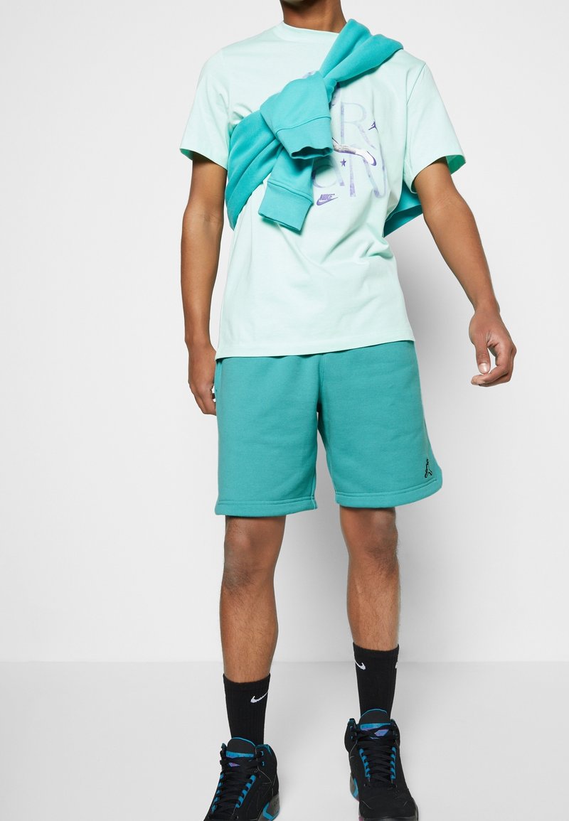 Homme portant un t-shirt vert menthe et un short sarcelle, avec un sweat sarcelle posé sur les épaules et des baskets noires avec des chaussettes noires.
