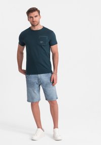 Camiseta verde oscuro con un bolsillo en el pecho, combinada con pantalones cortos de mezclilla azul claro y zapatillas blancas. Diseño simple, de mangas cortas y ajuste casual.