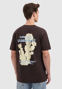 Young man with short brown hair wearing a dark brown t-shirt featuring a cactus graphic and text about Wasteland and southwestern US states.