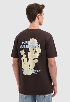 Jonge man met kort bruin haar, gekleed in een donkerbruine t-shirt met een cactusafbeelding en tekst over Wasteland en de zuidwestelijke staten van de Verenigde Staten.