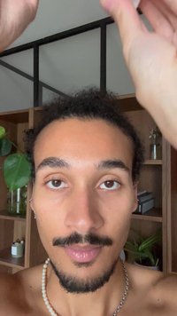 Mann mit lockigem Haar und Schnurrbart, trägt eine Perlenkette, vor hölzernen Regalen mit Pflanzen. Natürliches Licht, neutraler Hintergrund.