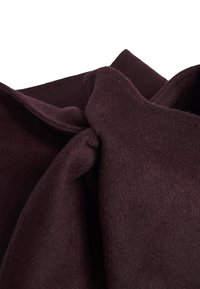 Tessuto di lana color bordeaux con una consistenza morbida, caratterizzato da un dettaglio a nodo attorcigliato. Il materiale appare spesso e accogliente, adatto per abbigliamento esterno.
