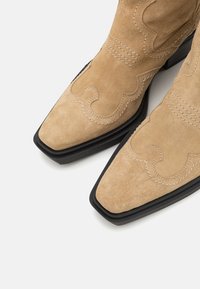 Botas de cowboy de ante marrón con punteras afiladas, costuras decorativas y una suela de goma negra a contraste. Detalles de diseño simples pero distintivos presentes.