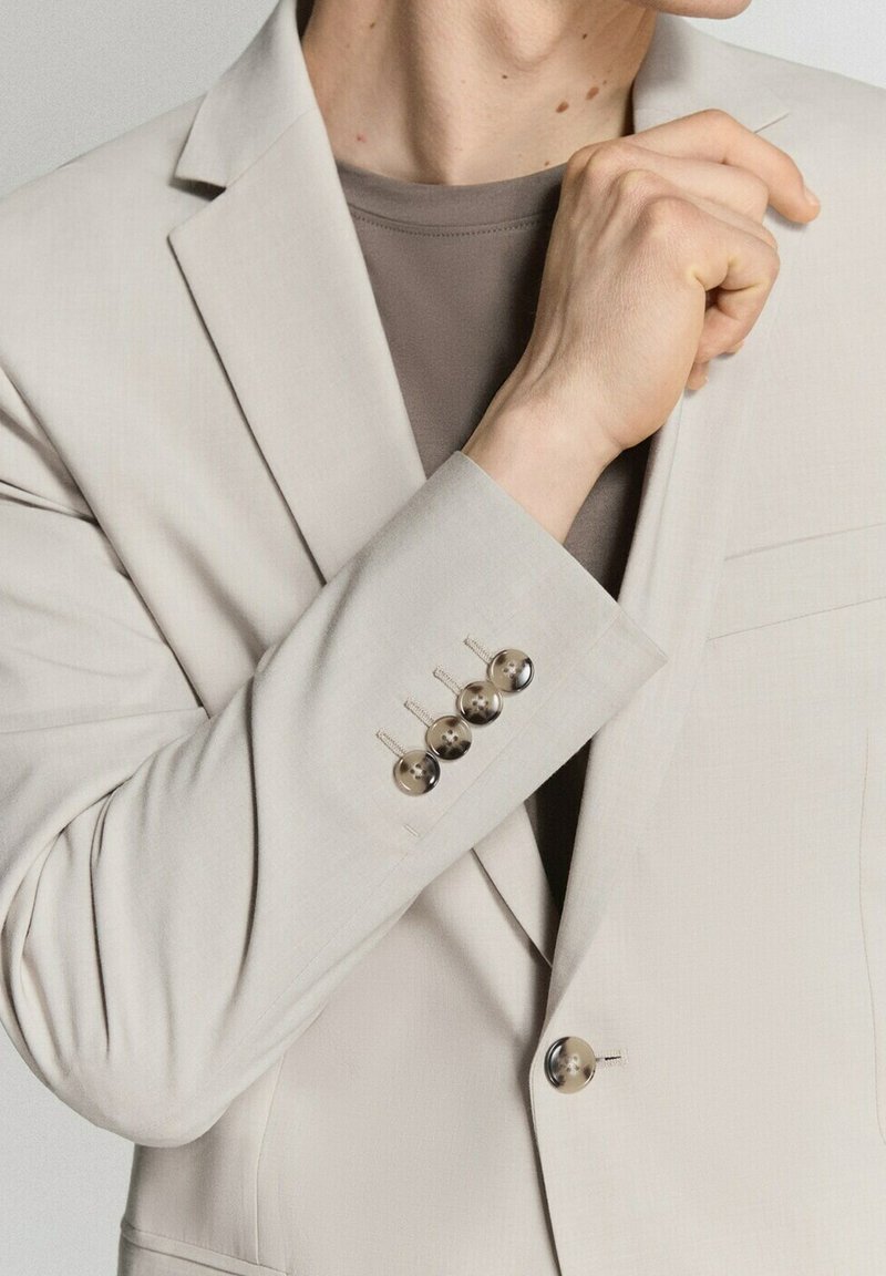 Persona ajustándose el cuello de un blazer beige con cuatro botones en la manga, vistiendo una camisa topo debajo.