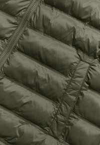 Maslinasto zelena puf jakna od tkanine s kvadratnom teksturom, s dijagonalnim šavovima i patentnim zatvaračem duž jednog ruba. Glatka i sjajna površina.