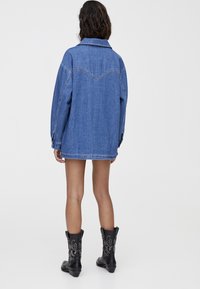 Persona con cabello oscuro y ondulado que lleva una chaqueta de mezclilla azul oversize y botas negras hasta la mitad de la pantorrilla con bordados blancos, de pie sobre un fondo blanco liso.