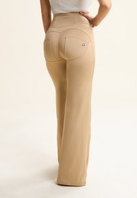 Pantaloni beige a vita alta con una vita aderente, parte frontale liscia e gambe svasate. Presentano due tasche posteriori con cuciture arcuate e un piccolo dettaglio del logo.