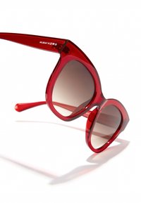 Piros túlméretezett napszemüveg fényes műanyag kerettel, átmenetes barna lencsékkel és elegáns kialakítással, amely egy logót tartalmaz az oldalán.