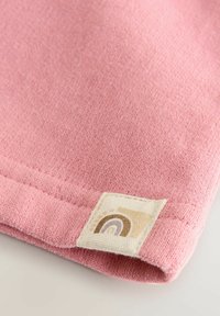 Nahaufnahme eines rosa Strickstoffs mit einem genähten beigen Etikett, das ein kleines Regenbogenmuster in Braun- und Cremetönen zeigt.