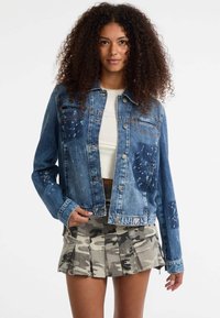 Giacca di denim con un lavaggio blu sbiadito, dettagli usurati, due tasche sul petto e hardware argentato, abbinata a una gonna plissettata camouflage.