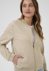 Veste bomber beige clair avec fermeture éclair à l'avant, poignets côtelés et poches latérales. Tissu lisse au design minimaliste.
