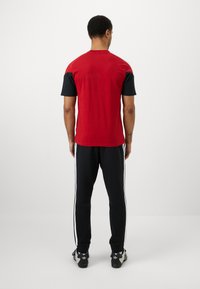Camisa vermelha de mangas curtas com mangas pretas, combinada com calças de jogging pretas com riscas brancas laterais. Ambas com um corte descontraído.