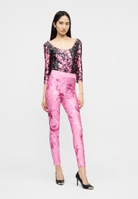 Μια γυναίκα στέκεται φορώντας ένα μπλουζάκι με μαύρο και ροζ μοτίβο λεπιών και λαμπερές ροζ κολάν με σχέδιο, συνοδευόμενα από μαύρες ψηλοτάκουνες γόβες πάνω σε λευκό φόντο.