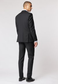 Schwarzer Anzug mit schlanker Passform. Mit strukturierten Schultern, Schlitz hinten und Zweiknopfverschluss. Getragen mit schwarzen Schuhen und weißem Hemd.