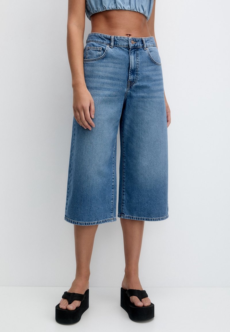 Culottes de denim de color azul claro con una cintura alta, piernas anchas y dobladillo sin terminar. Combinados con sandalias de plataforma negras y gruesas y una blusa corta.