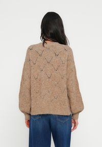 Pull marron tricoté avec un motif en treillis diamant, coupe ample et manches bouffantes, associé à un jean bleu à jambes larges.