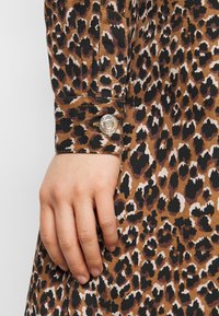 Leopardmönstrad klänning i bruna nyanser med svarta accenter. Har en knäppt manschett med en metallisk knappdetalj, som visar upp ett texturerat tyg.