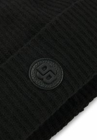 Berretto di maglia nera con texture a coste e un patch circolare in pelle impresso con il logo 'PB' sul davanti.