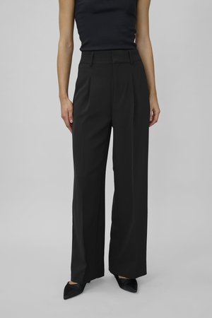 Schwarze, hochtaillierte, weit geschnittene Hose aus einem glatten Stoff, mit vorderen Falten und Gürtelschlaufen, kombiniert mit spitzen schwarzen Schuhen.
