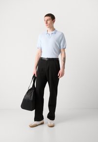  világoskék kötött pólóing gallérral, fekete nadrággal és fehér sportcipővel. Egy fekete nylon válltáskát hord.