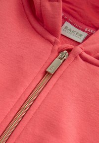 Rozā džemperis ar gludu virsmu, ar zeltītu rāvējslēdzēju un kapuci. Uz birkas rakstīts "Baker by Ted Baker." Tuva fokusa uzmanība uz rāvējslēdzēja detaļu.