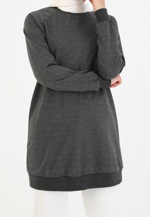 Osoba ubrana w ciemnoszary długi tunikę z prążkowanymi mankietami i dołem, w połączeniu z białymi spodniami i białą chustą na głowę.