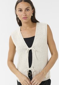 Knit sleeveless vest in off-white with a deep V-neck and front tie closure. Worn over a black top, featuring a textured fabric.