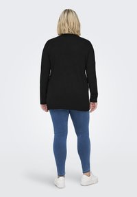 Zwarte gebreide trui met lange mouwen, iets langere achterkant, gecombineerd met blauwe skinny jeans en witte sneakers. Achteraanzicht weergegeven.