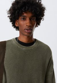 Knit sweater in olive green with a textured pattern, wide crew neck, and black undershirt visible. Brown shoulder accents complete the look.