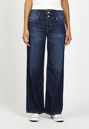 Frau trägt dunkelblaue Jeans mit weitem Bein und weiße Sneaker, steht vor einem einfarbigen hellen Hintergrund.