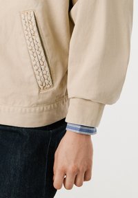 Krem bomber jakna z teksturiranim pletenim detajlom na žepu, rebrastimi manšeti in sproščenim krojem. Nosi se preko modro prugaste srajce.