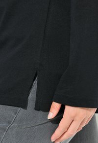 Schwarzes Langarmshirt mit einer weichen Textur, das einen seitlichen Schlitz am Saum und kontrastierenden grauen Unterstoff aufweist. Nahaufnahme des Shirts.