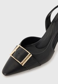 Juodas odinis slingback aukštakulnis batelis su smailiu toe, puikuojantis auksiniu sagtimi ir įspaustu logotipu ant dirželio. Lygi tekstūra, elegantiškas dizainas.