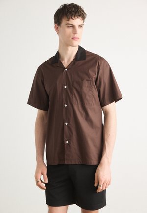 Brown short-sleeve shirt with a black collar, white buttons, and a chest pocket. The fabric appears smooth and slightly glossy.