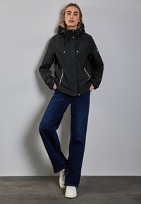 Svart vattentät jacka med huva, silverfärgade dragkedjor och justerbar snörning. Matchas med mörkblå utsvängda jeans och vita sneakers.