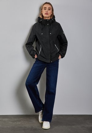 Sort vandtæt jakke med hætte, sølvfarvede lynlåse og justerbar snor. Sammen med mørkeblå slå-ud jeans og hvide sneakers.