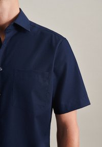 Camicia a maniche corte blu navy in tessuto liscio, con colletto classico e una tasca sul petto. Bottoni vicino al colletto.