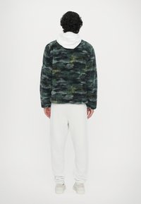 Fleece jacket in green and black camo pattern, white hoodie beneath, paired with white sweatpants and beige sneakers.