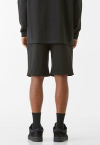 Crna majica dugih rukava u paru s crnim šortsevima, oboje od glatke tkanine. Crne čarape i tenisice upotpunjuju odjevnu kombinaciju, prikazano s leđa.