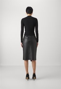 Eine Frau, die mit dem Rücken zur Kamera steht, trägt ein schwarzes Langarmoberteil, einen schwarzen Lederrock bis zum Knie, schwarze High Heels und goldene Creolen.