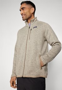 Giacca felpata beige con zip, colletto alto, tessuto texturizzato, tasche laterali e rifiniture beige a contrasto. Logo sul petto sinistro.
