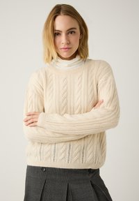 Cremefarbener Kabelstrickpullover mit gerippten Rändern, lockerer Passform und strukturierten Mustern. Kombiniert mit einem dunklen Plisseerock.